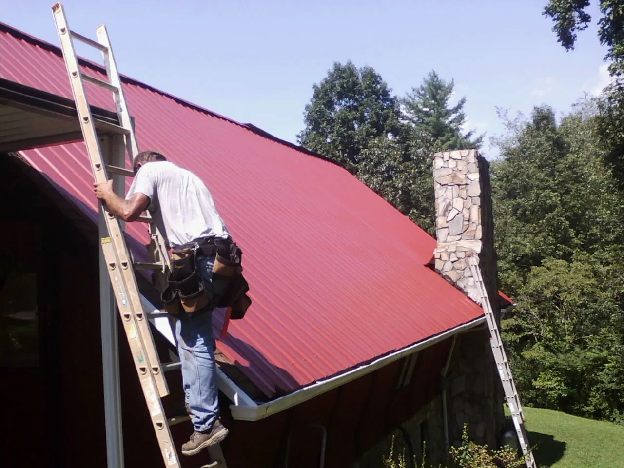 Roofer on a ladder beside a red metal roof and stone chimney — Soffit & Fascia Repair by Roofing Brothers in Schellsburg